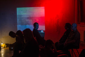 Seven people sitting in chairs seen mostly in silhouette in a dark room with a red light and projection of a train on the wall.