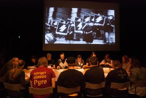 A group of people sitting around a table reading from papers. Bheind them a projection of a photograph of Hitler Jugend from Nazi Germany.